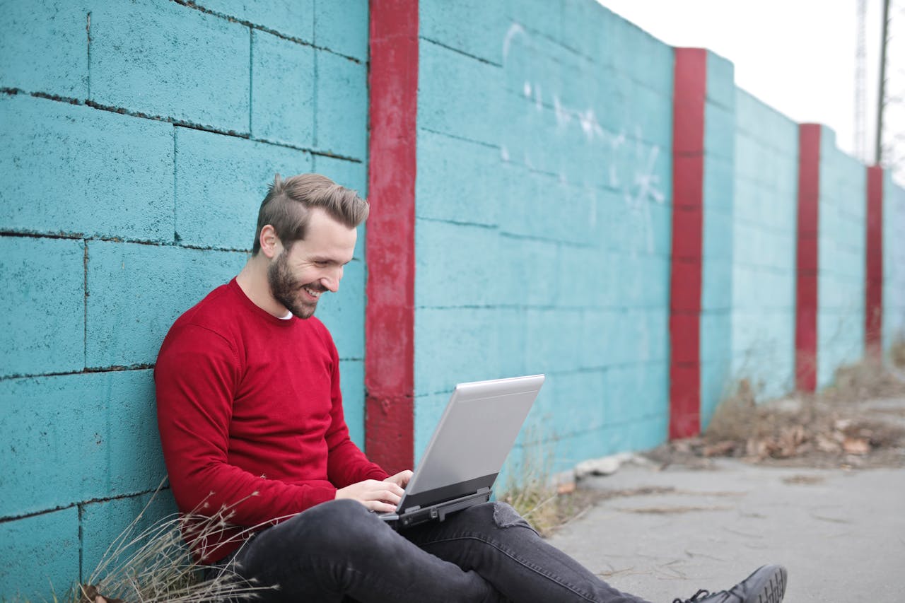 Home A man smiling and using a laptop outdoors against a vibrant blue wall, representing remote work.
